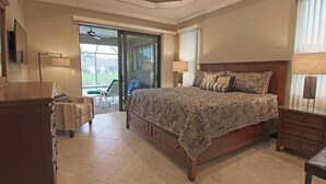 A bedroom with a large bed, a dresser, a nightstand, and a chair. There is a sliding glass door leading to a balcony.