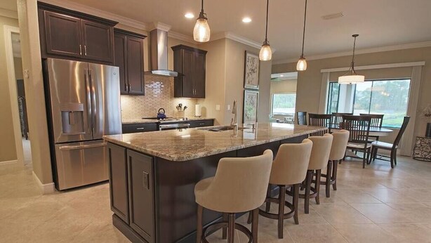 A modern kitchen with dark cabinetry, stainless steel appliances, a granite island with bar stools, pendant lighting, and a large window providing natural light.