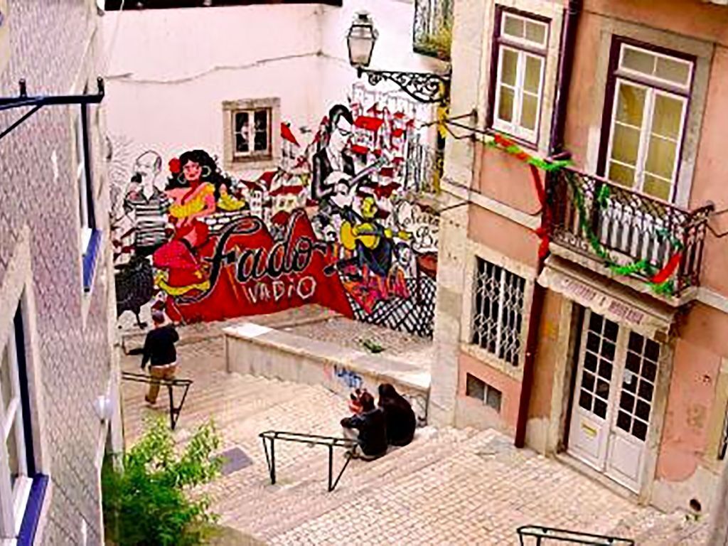 A narrow street with a mural on a building, a staircase, and a balcony with railings.