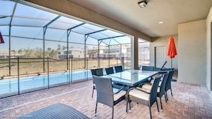 A covered patio with a glass table and chairs, a brick floor, and a view of a swimming pool and houses.