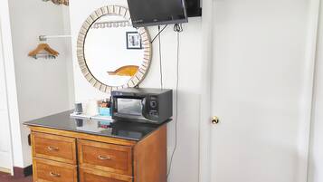 Une pièce comprenant une commode en bois, un four à micro-ondes, un téléviseur fixé au mur, une porte avec une poignée de porte en laiton et un miroir rond orné d’un motif floral.