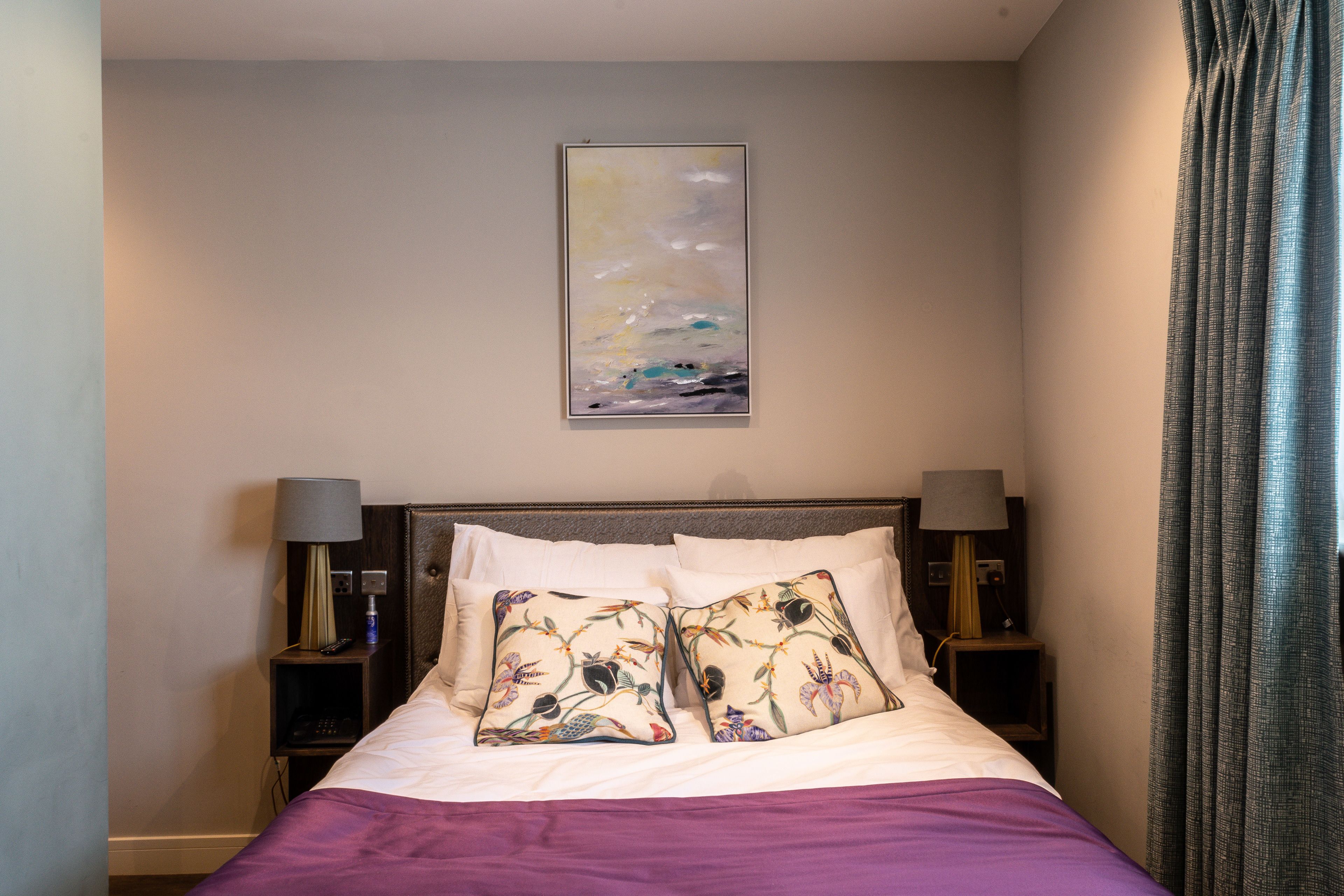 Una cama bien hecha con almohadones florales, un cuadro en la pared encima y dos lámparas de noche.