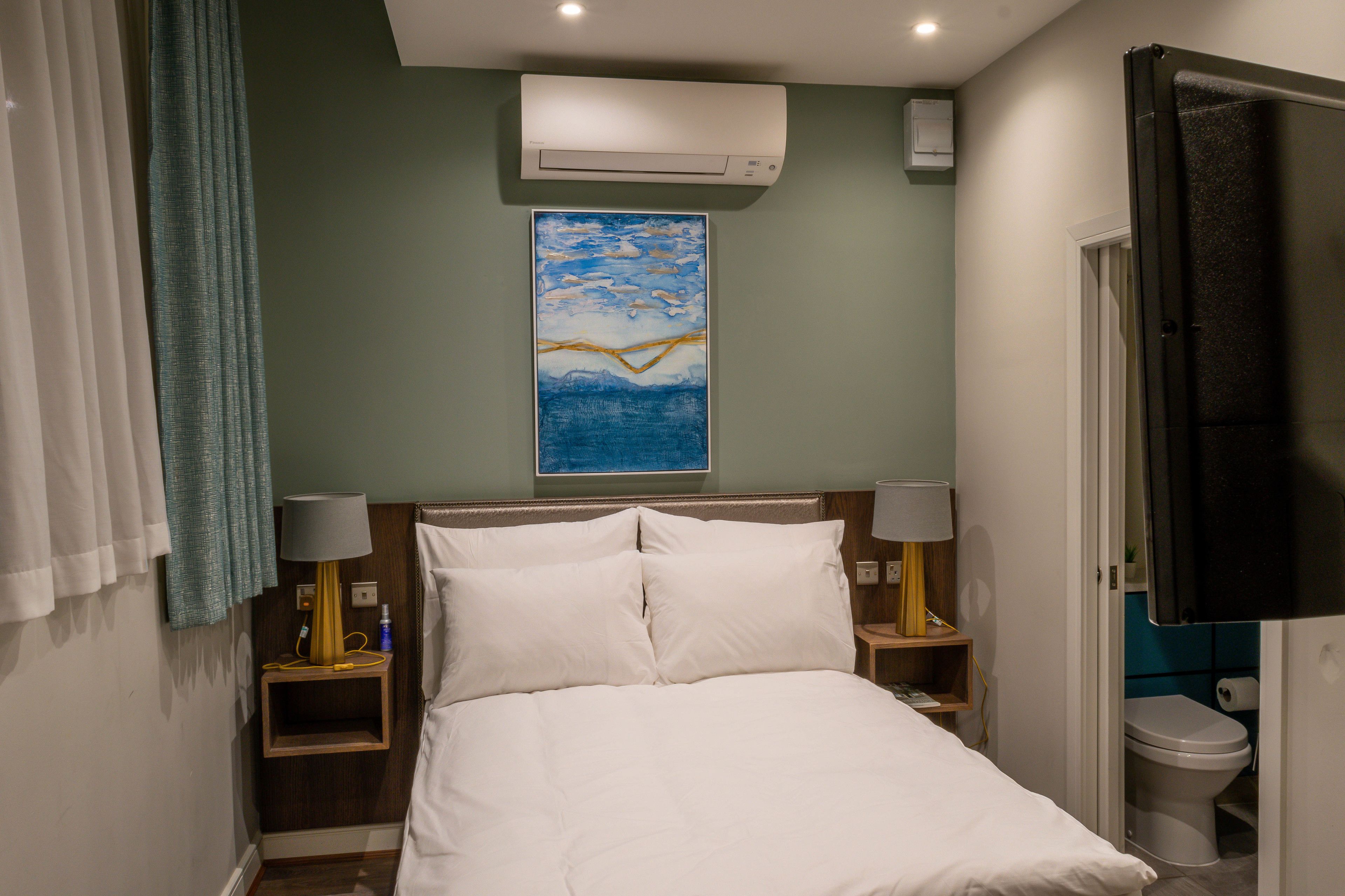 Hotelkamer met een bed, twee bedlampen, een airconditioner, een schilderij aan de muur, een badkamer met toilet en een aan de muur bevestigde televisie.