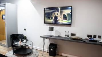 Una sala moderna con un televisor de pantalla plana montado en la pared, una mesa de centro redonda de vidrio, una silla negra y un pequeño cubo de basura.