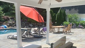 A covered patio area with a pool, lounge chairs, a wooden table, and an umbrella. - A dream summer rental 30 miles from New York City in picturesque Bedford, NY! (Bedford)