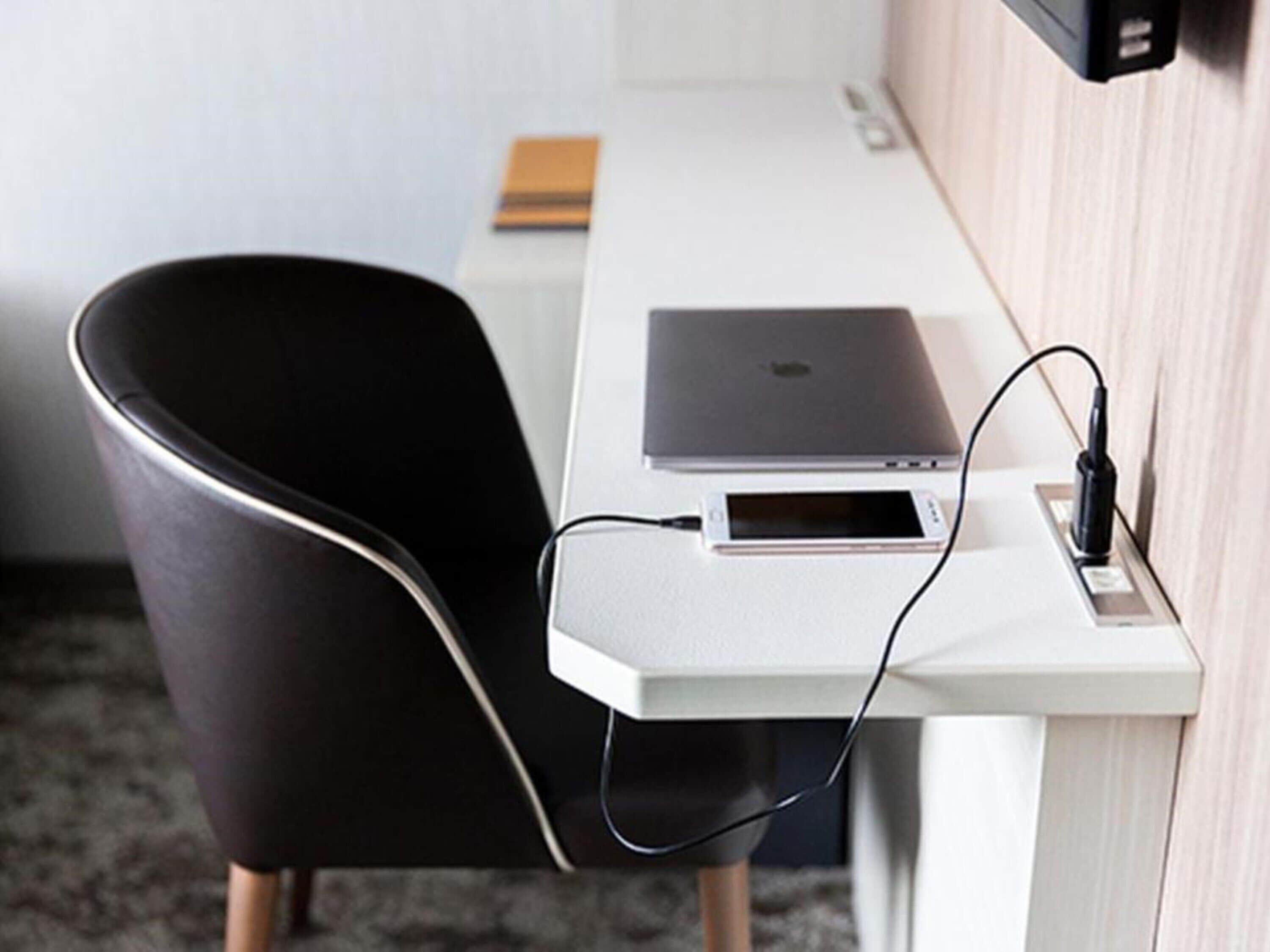 A modern office chair, a white desk, and a laptop with a smartphone connected to a power source.