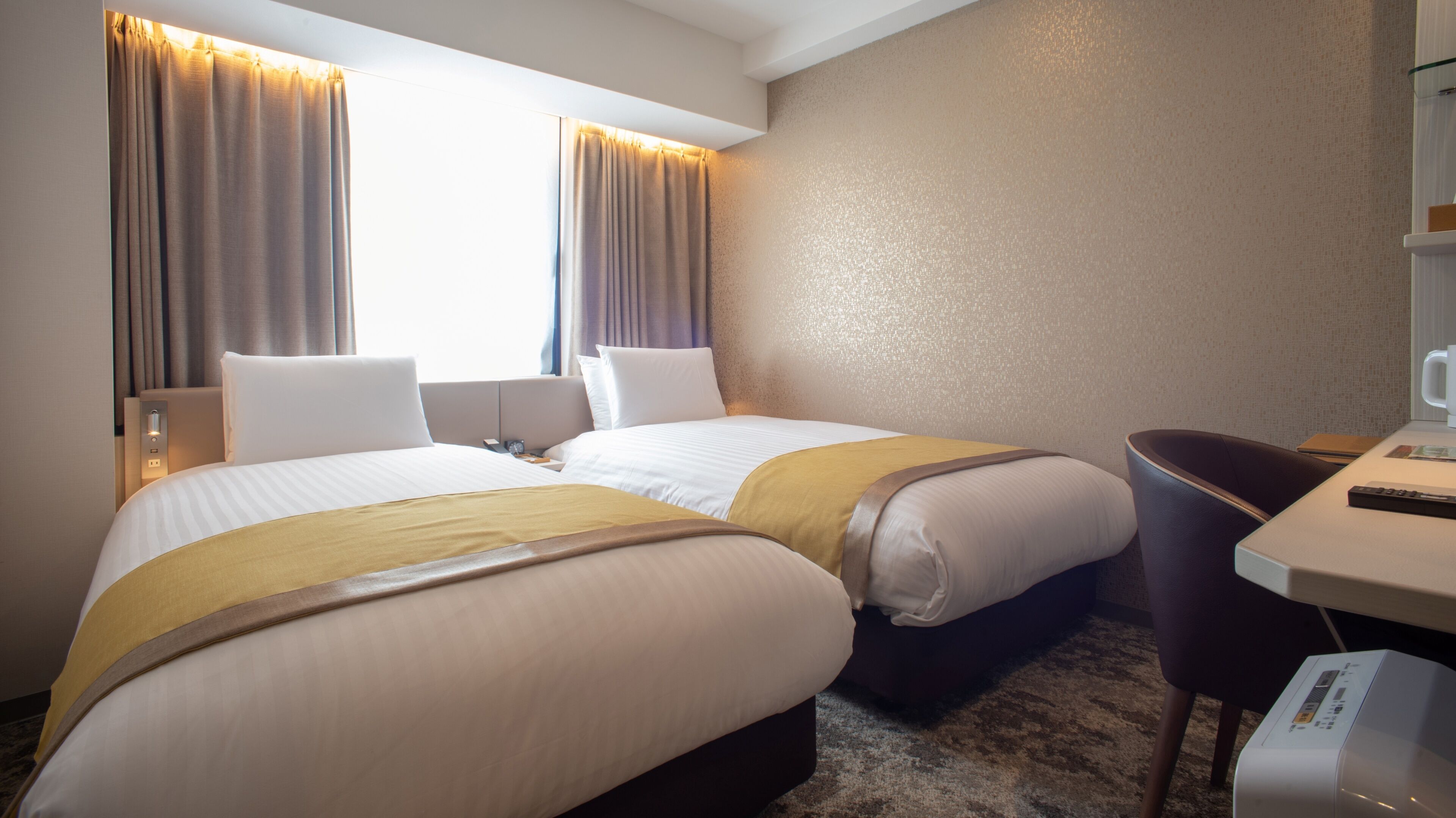 Quarto de hotel com duas camas, uma escrivaninha, uma cadeira, uma máquina de café e uma janela com cortinas.