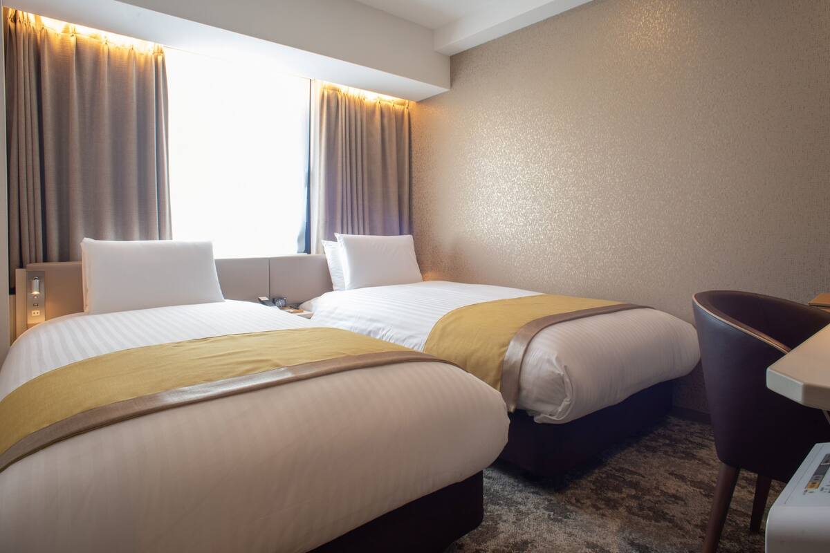 Quarto de hotel com duas camas, uma escrivaninha, uma cadeira, uma máquina de café e uma janela com cortinas.