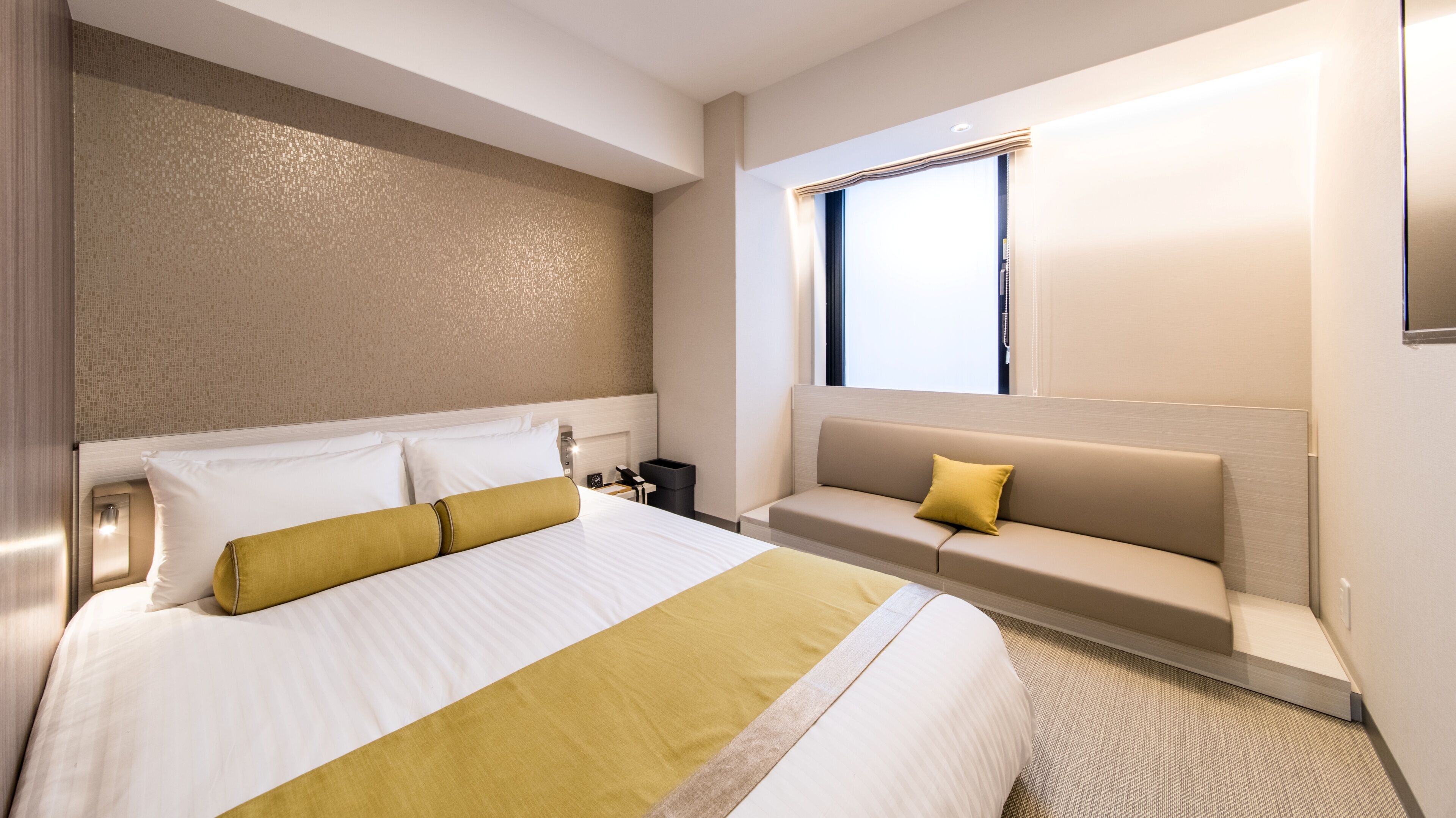 Quarto de hotel com uma cama grande, um sofá, uma mesinha de cabeceira e uma janela.