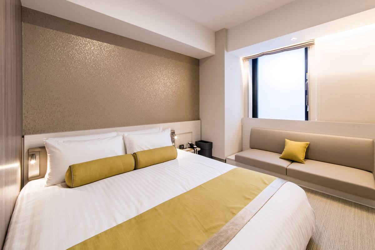 Quarto de hotel com uma cama grande, um sofá, uma mesinha de cabeceira e uma janela.