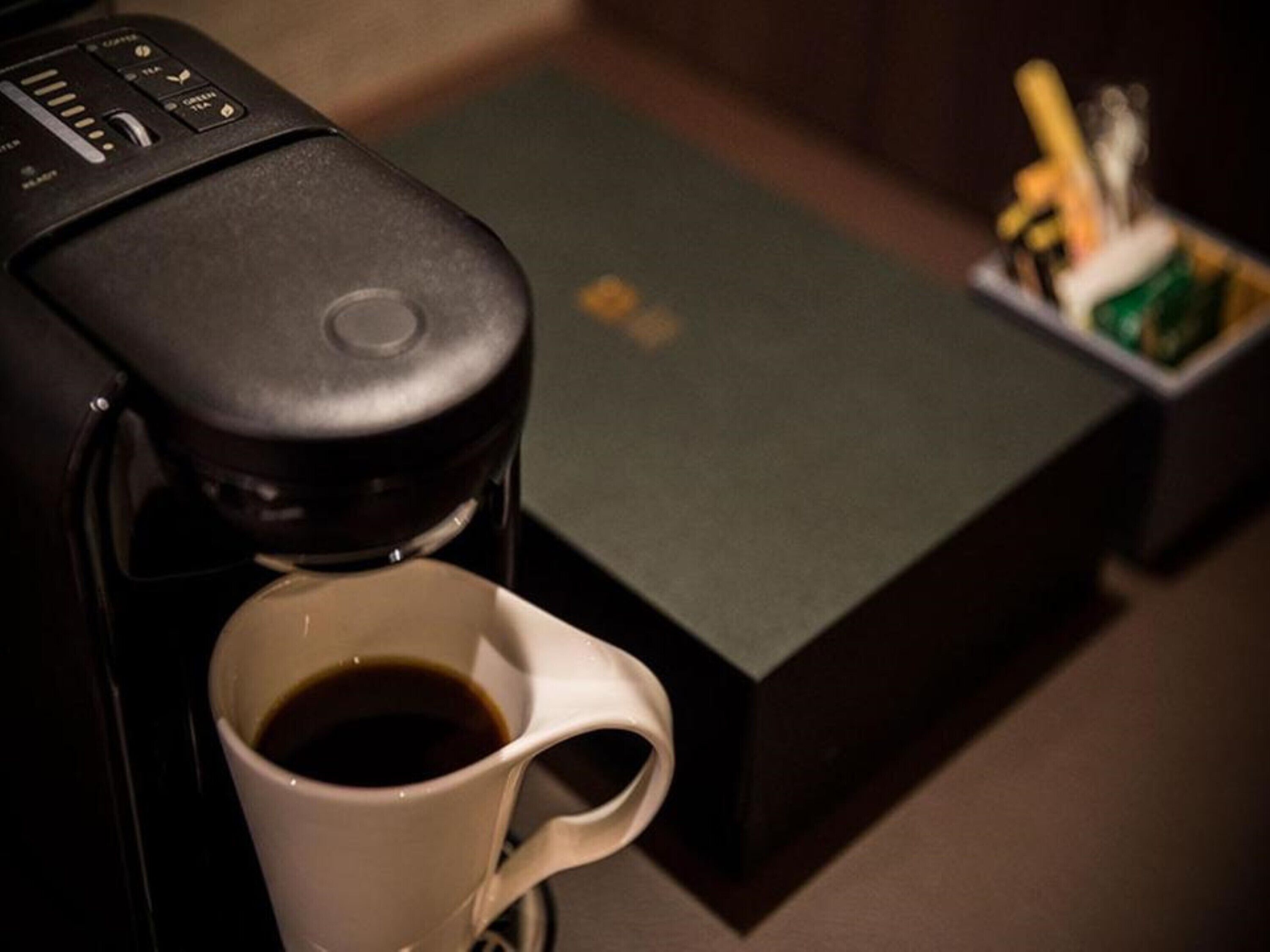 A coffee machine dispensing coffee into a cup, with a box and a pen holder in the background.
