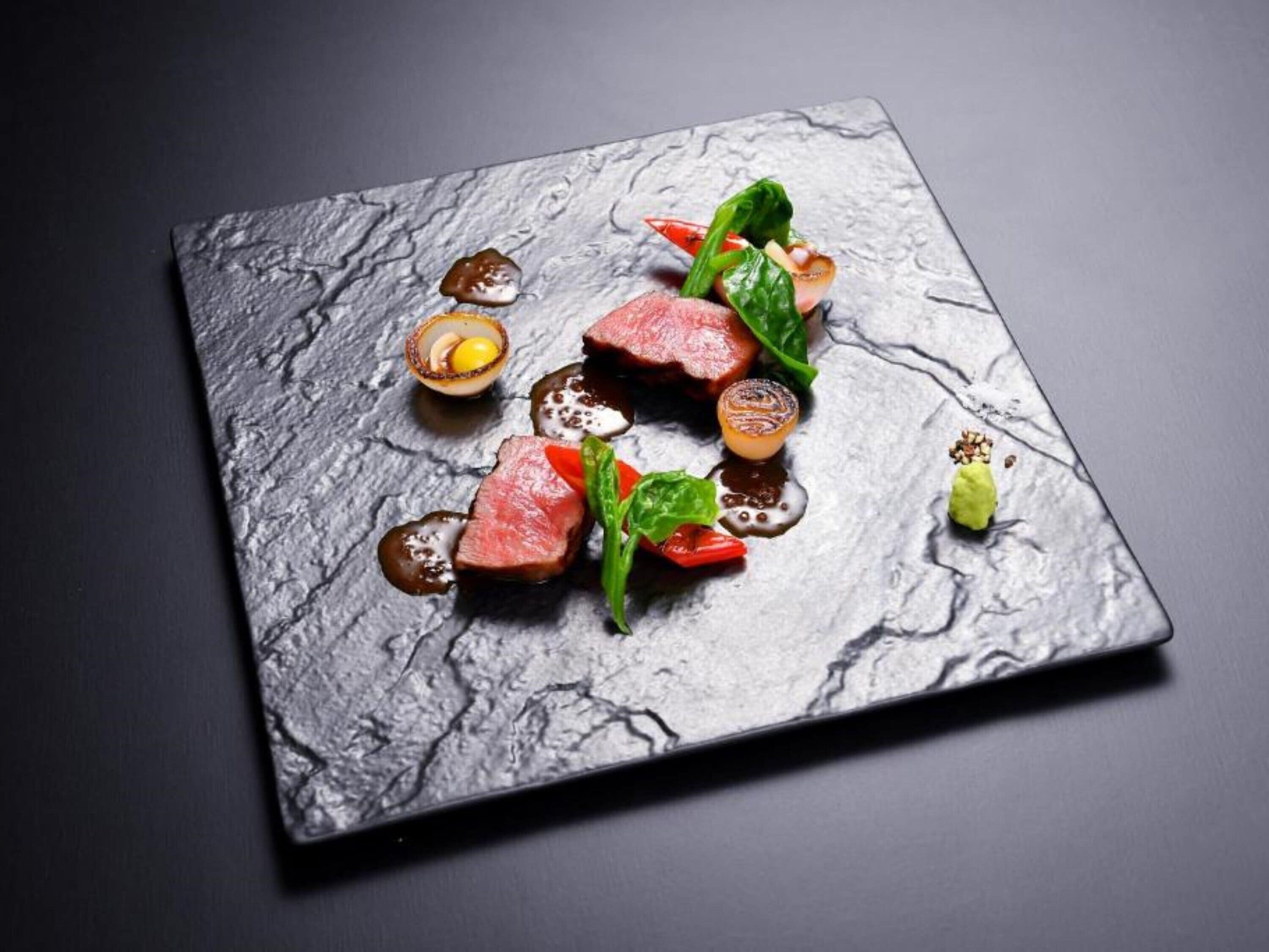 A square plate with a textured surface, containing a dish with meat, vegetables, and a sauce.