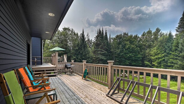 Una terraza de madera con mobiliario exterior, una parrilla y vistas a un área boscosa.