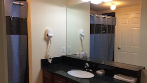 Un baño con encimera negra, lavabo blanco y un espejo grande. Hay dos secadores de pelo fijados en la pared y una toalla doblada sobre la encimera.