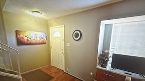 Una sala de estar con piso de madera, una puerta, un reloj, un cuadro, una ventana con persianas y un mueble para televisor.