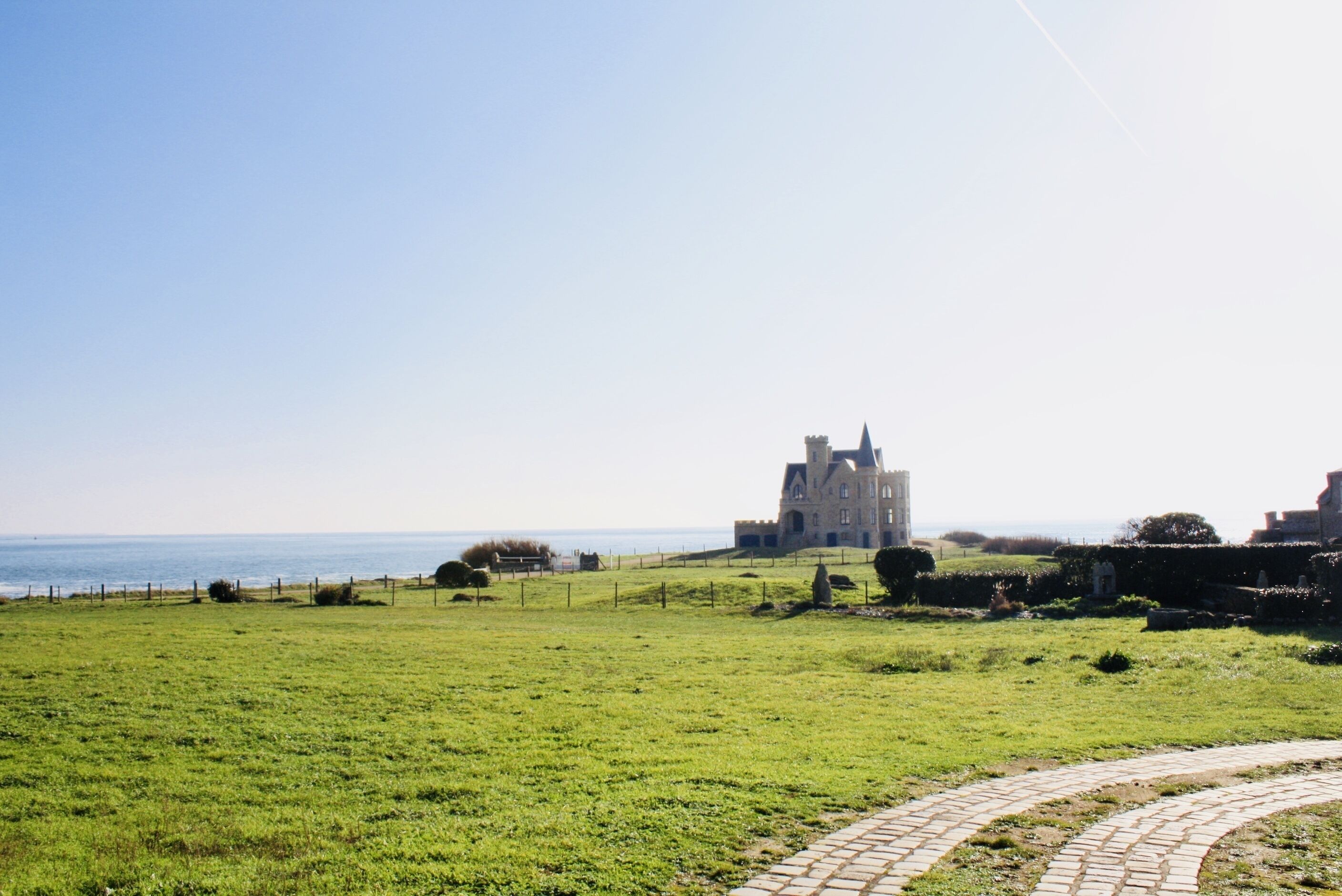 A grassy field with a stone pathway leading to a historic building by the sea.