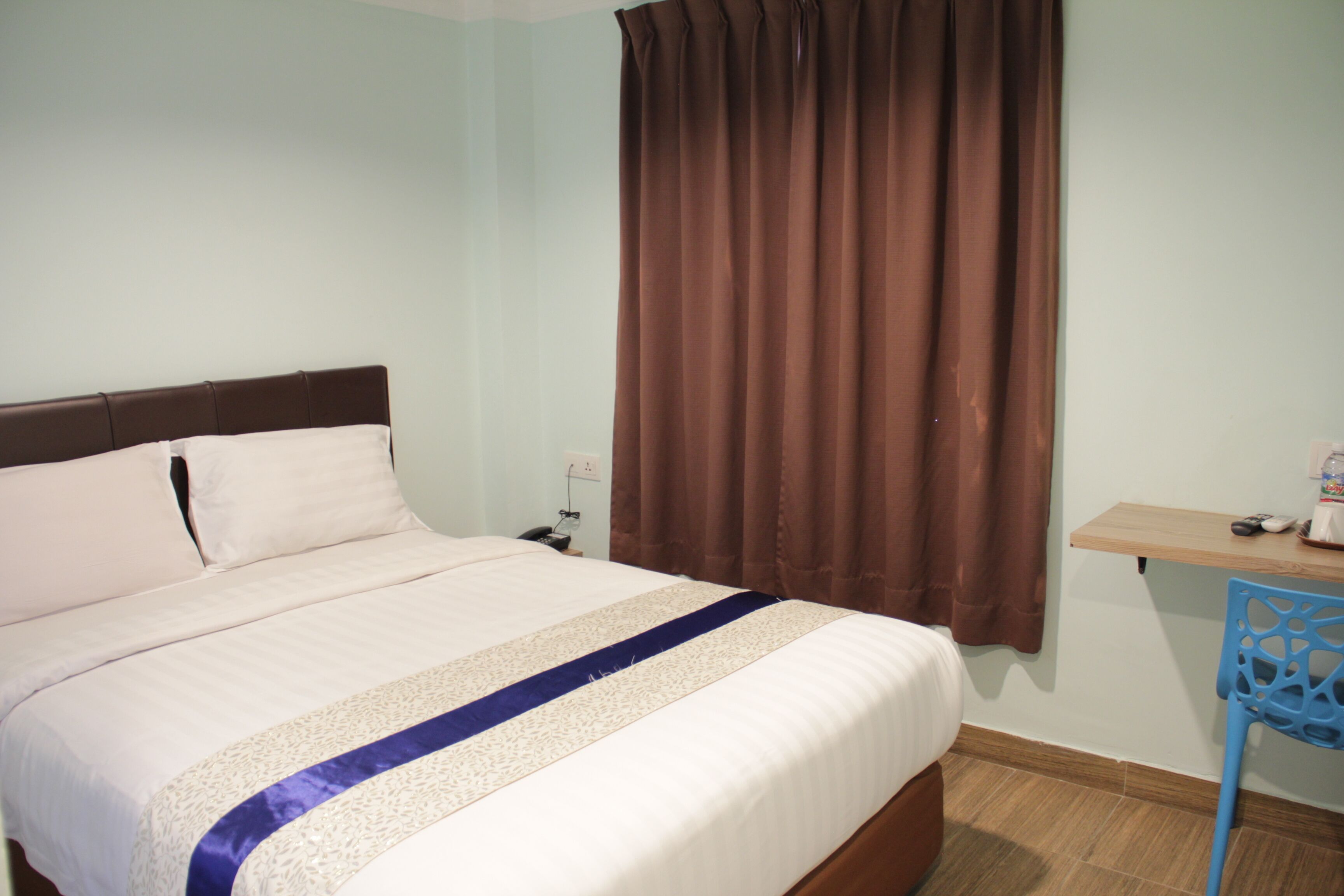 Uma cama bem arrumada com edredom branco e azul, cortina marrom e uma cadeira azul em um quarto com paredes em tons claros e chão de madeira.