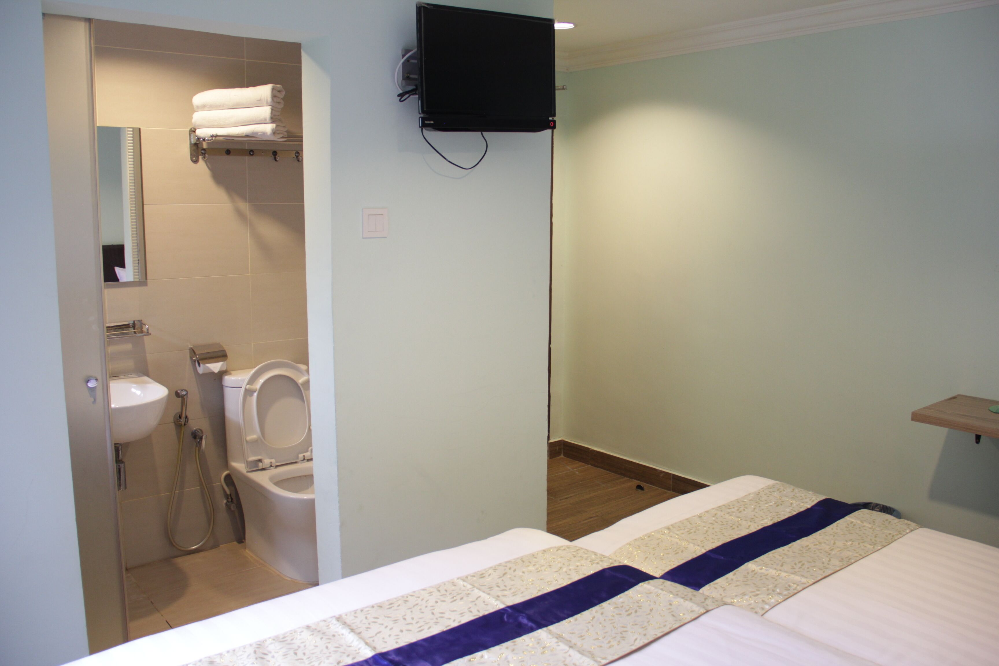 A bathroom with a toilet, sink, and shower. There is a bed with a white and purple bedspread. A flat-screen TV is mounted on the wall.