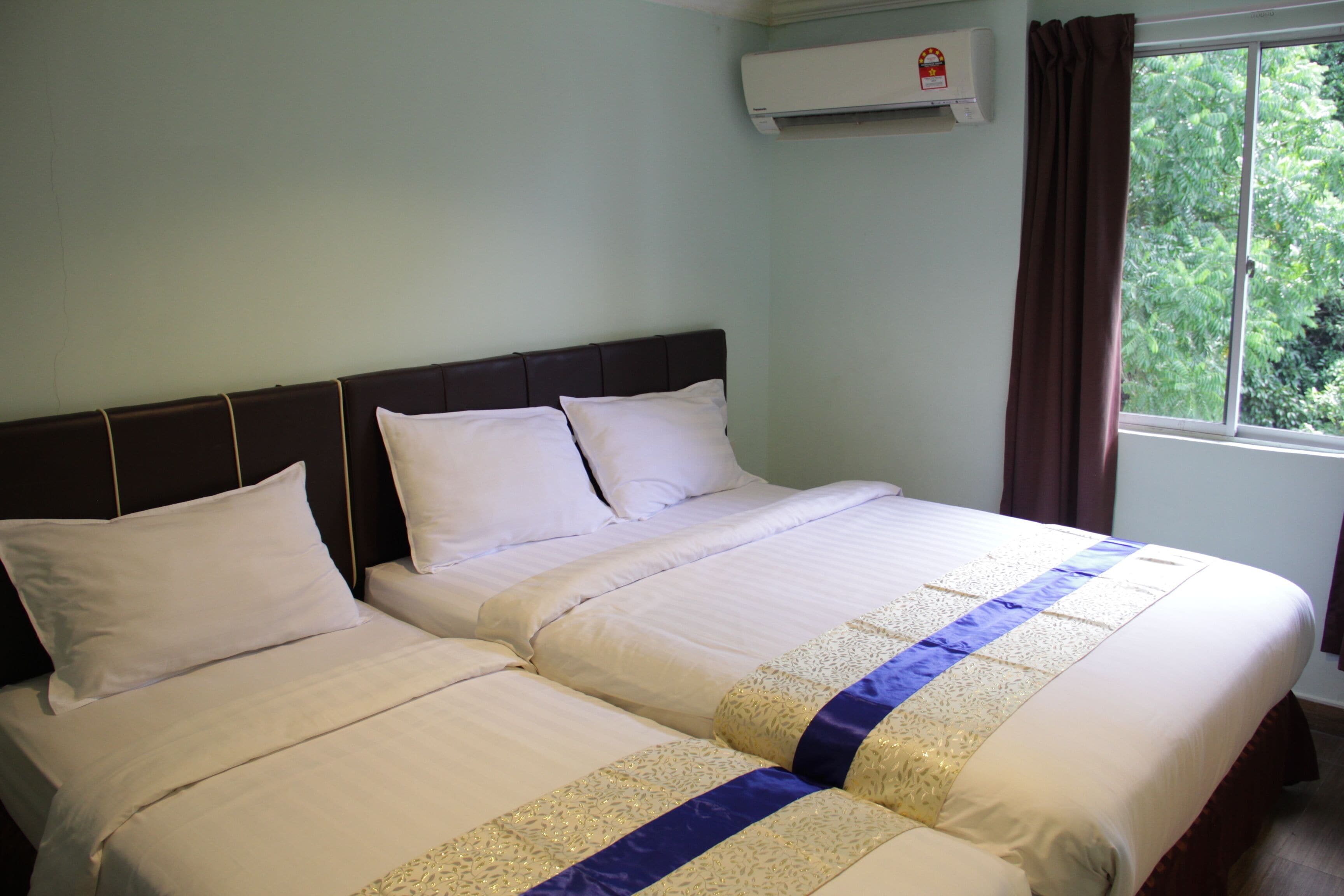 A double bed with white linens and a blue and gold decorative strip, a dark headboard, and a window with a brown curtain.