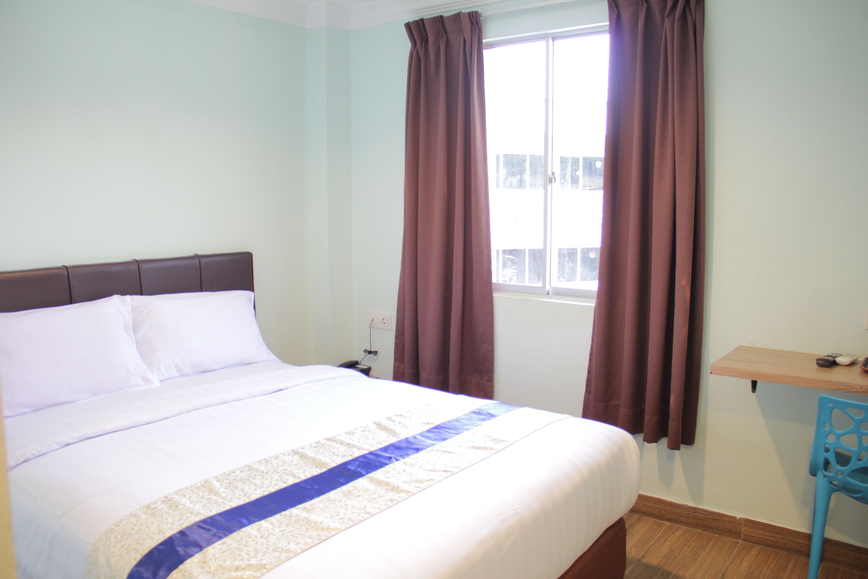 A neatly made bed with a white and blue bedspread, a wooden headboard, and a window with brown curtains.