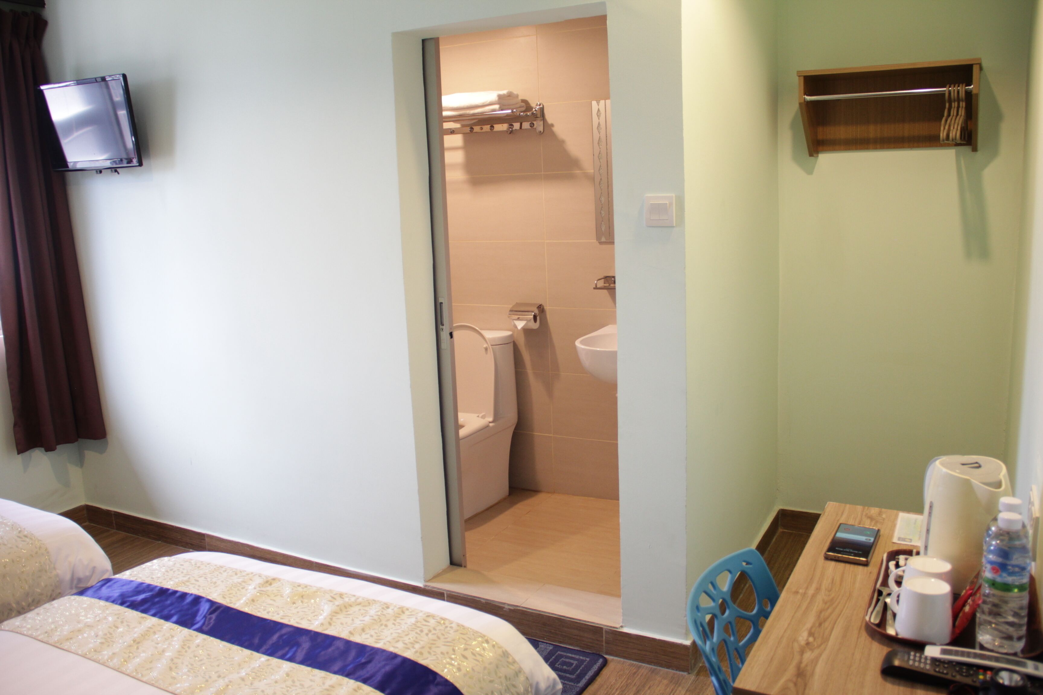 A hotel room with a bed, a wooden table with toiletries, a TV mounted on the wall, and a bathroom visible through an open door.
