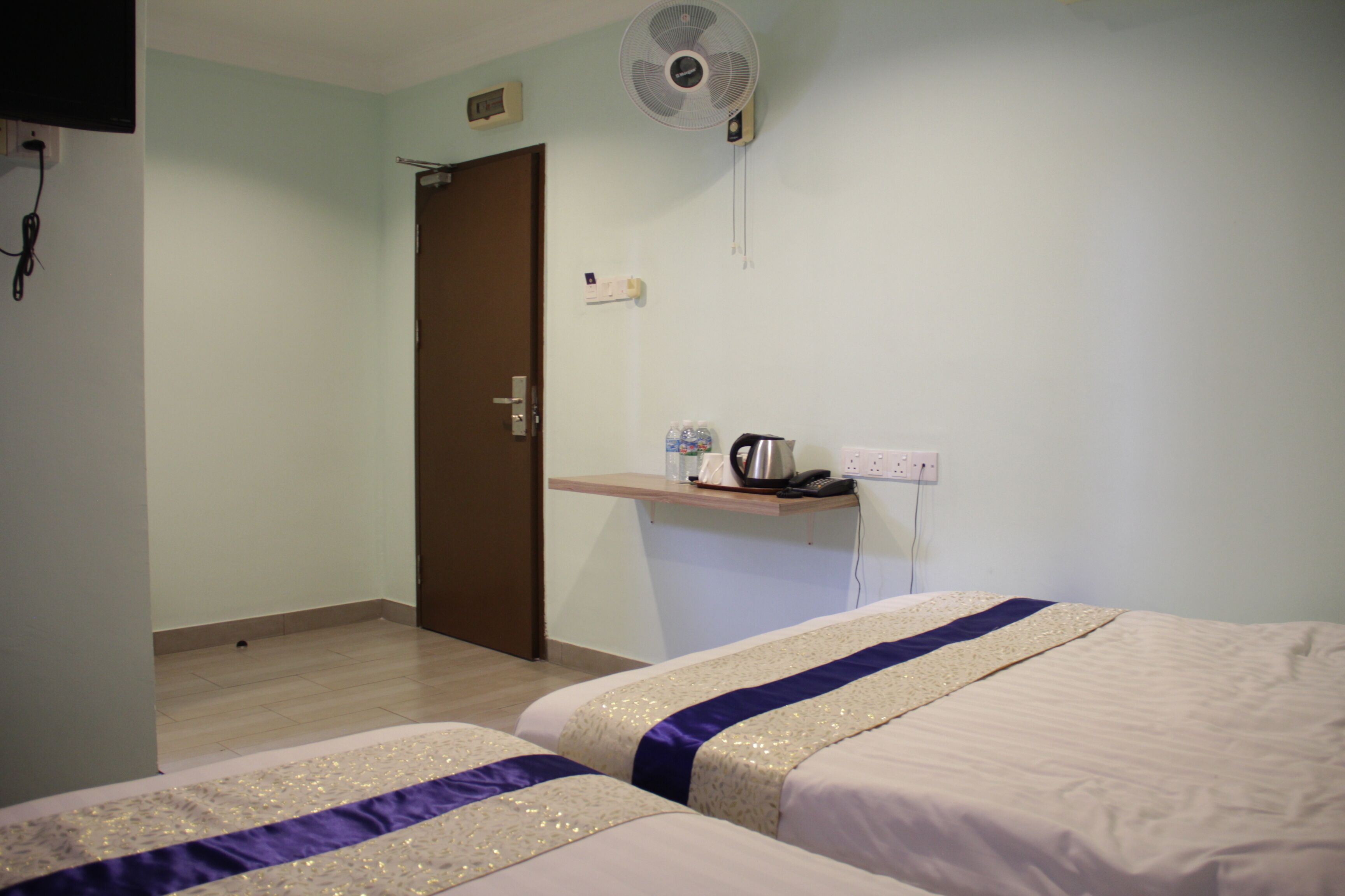 A hotel room with two beds, a small desk with a kettle, a fan, and a door.
