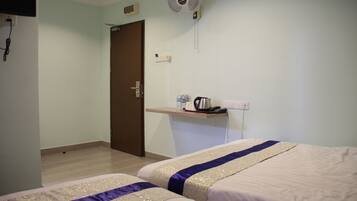 A hotel room with two beds, a small desk with a kettle, a fan, and a door.
