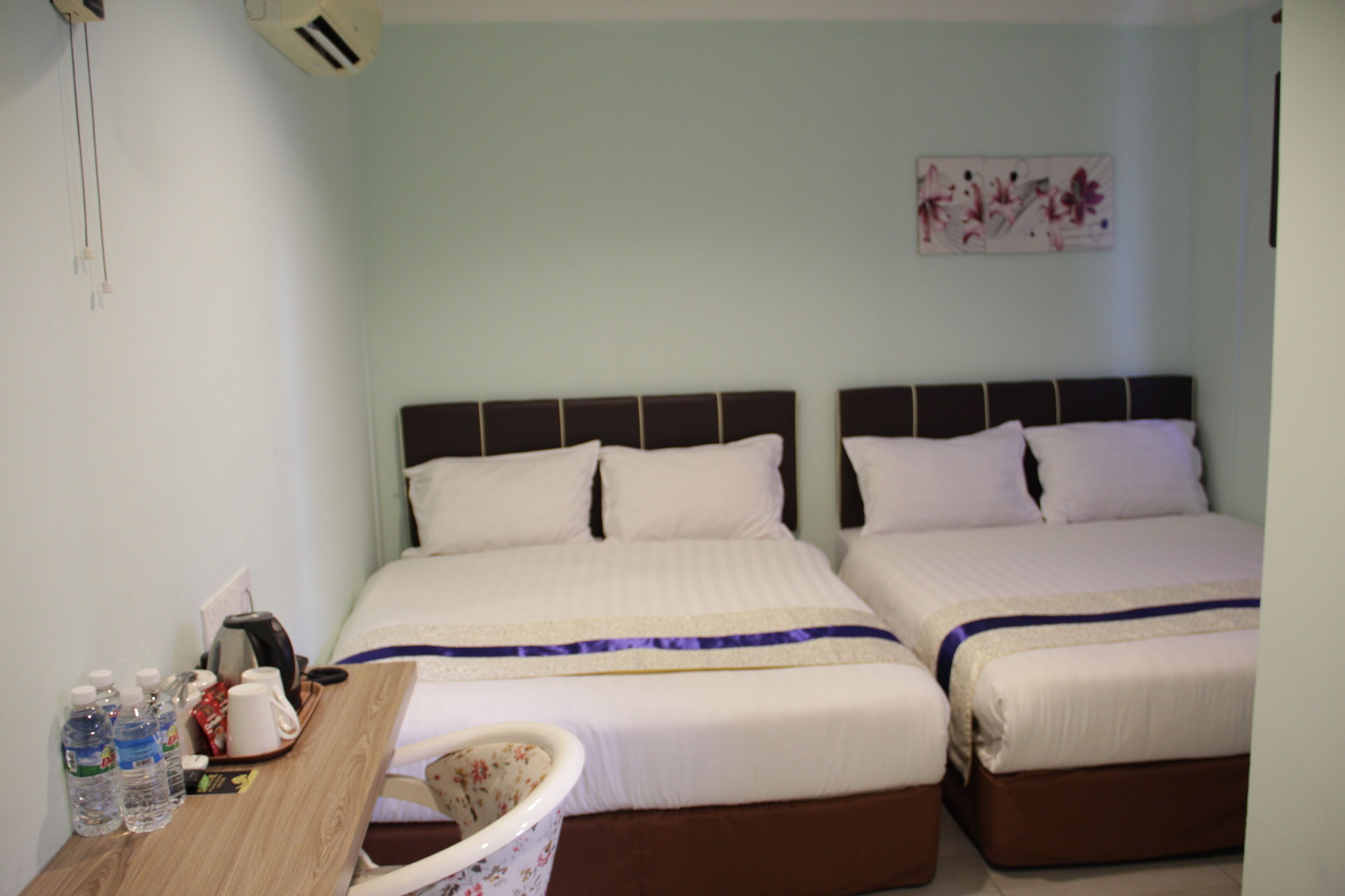 A hotel room with two beds, a wooden desk with a chair, and a small table with bottles and a kettle.