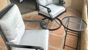 Un balcon avec deux fauteuils rembourrés et une petite table ronde, un lit avec une literie blanche, et une porte-fenêtre coulissante donnant sur une pièce.