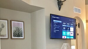 Un salon avec un téléviseur à écran plat fixé au mur, affichant un écran d’accueil, une décoration en forme de crâne de vache, des reproductions botaniques encadrées, un fauteuil à motifs et un meuble en bois.