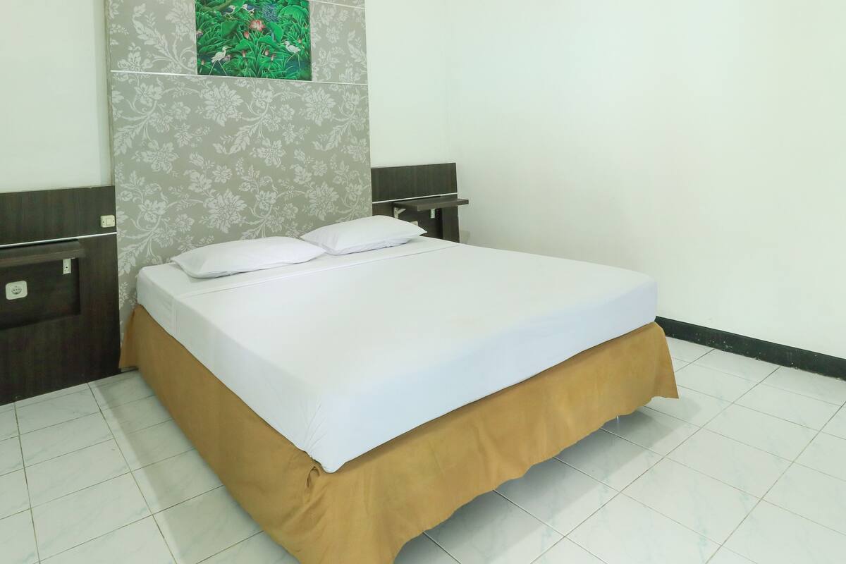 Cama bem arrumada com colchão branco e saia de cama marrom, mesa de cabeceira com abajur, e parede com padrão floral.