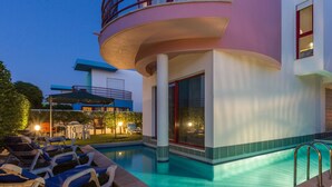 A swimming pool area with lounge chairs, a covered seating area, and a building with a balcony.