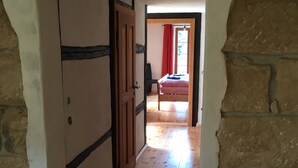A hallway with wooden flooring leading to a bedroom with a bed and a window with red curtains.