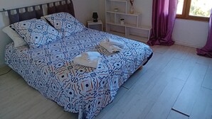 Una cama bien hecha con ropa de cama estampada, una mesita de noche con lámpara, una estantería con adornos y una ventana con cortinas.