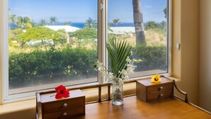 A room with a large window offering a view of the ocean and palm trees. There is a wooden table with a vase of flowers and a small wooden cabinet. - Kumulani I-1: Breathtaking Ocean Views w/ Wrap Around Lanai, Pool & AC (Kamuela)