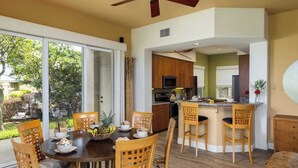 A modern kitchen with wooden cabinets, a central island, and a dining area with a round table and chairs. - Kumulani I-1: Breathtaking Ocean Views w/ Wrap Around Lanai, Pool & AC (Kamuela)