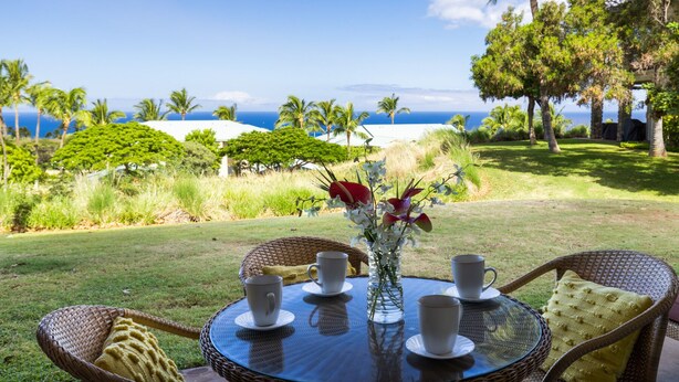 A round table with four white cups and saucers, a vase with flowers, and wicker chairs with cushions. - Kumulani I-1: Breathtaking Ocean Views w/ Wrap Around Lanai, Pool & AC (Kamuela)