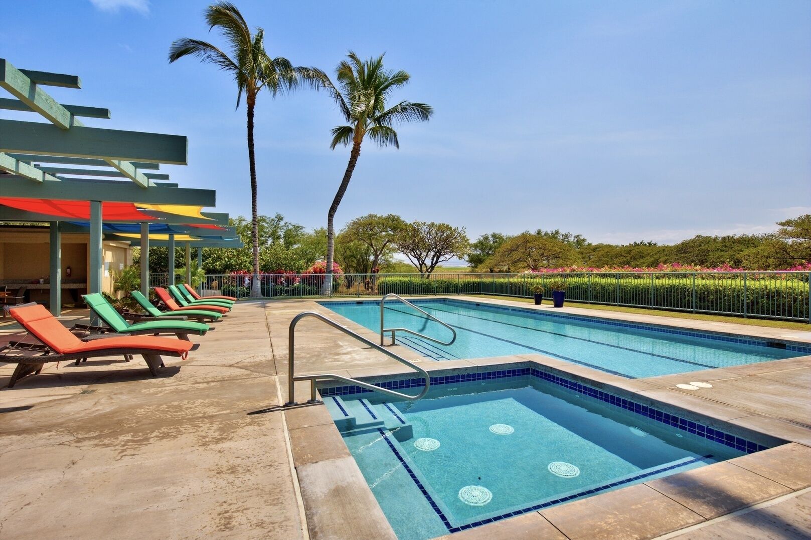 Une piscine avec une terrasse carrelée, des chaises longues, des palmiers et un espace abrité surmonté d’un toit aux couleurs vives.