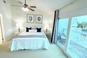 Une chambre à coucher avec un lit, deux tables de chevet avec des lampes, un ventilateur de plafond et une grande fenêtre donnant sur un balcon.