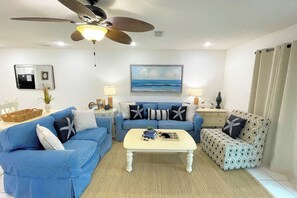 Ein Wohnzimmer mit Deckenventilator, Couchtisch, blauem Sofa, einem gemusterten Sessel und einem Meeresbild.