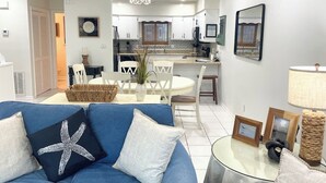 A modern living room with a blue sofa, a glass coffee table, and a dining area with white chairs. - Gulf Views · Steps to Beach & Beachfront Pool (Port St. Joe)