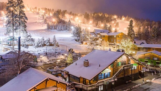 A snow-covered lodge with multiple buildings, a ski slope in the background, and a view of the surrounding forest.