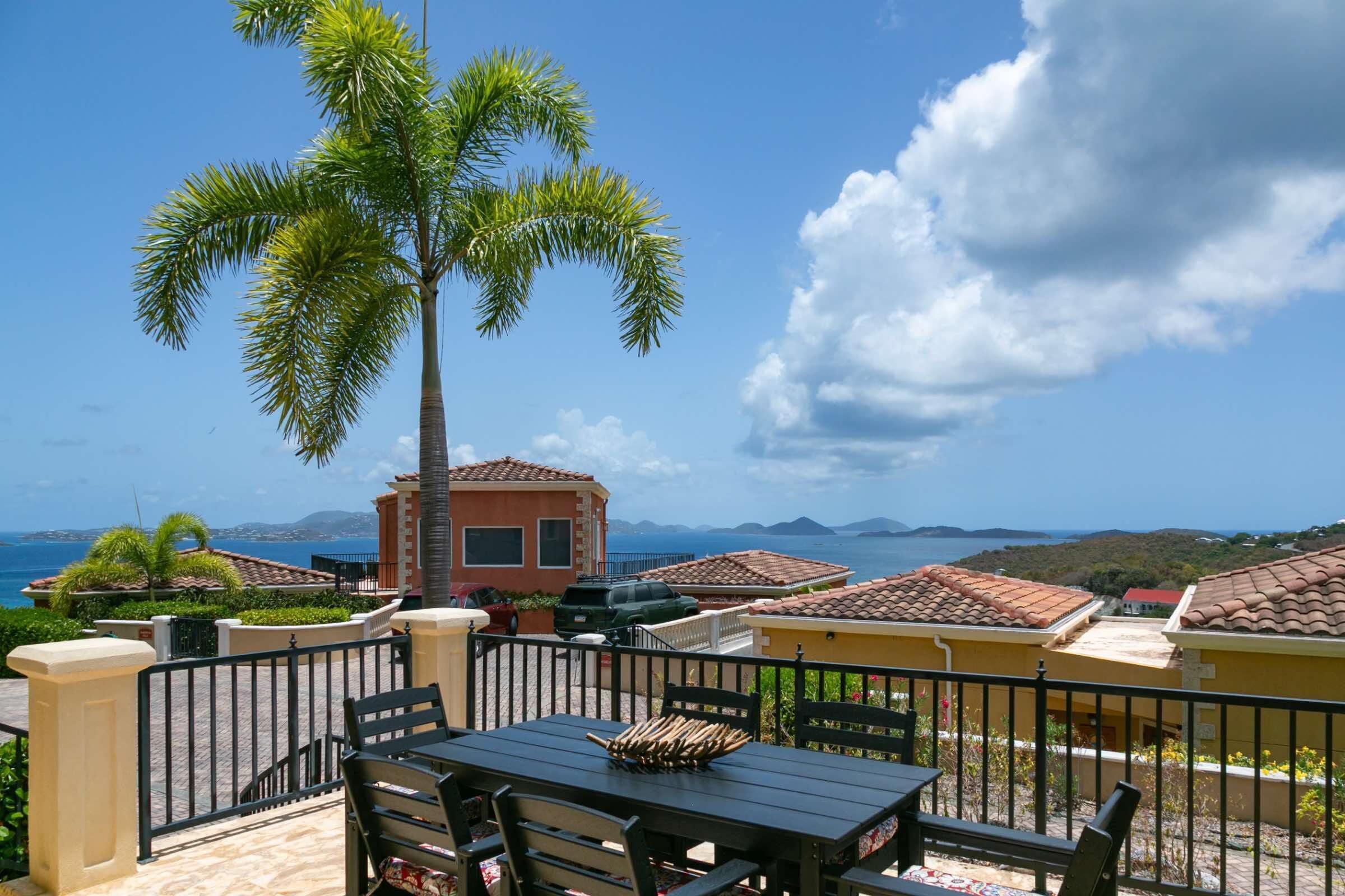 A patio with a table and chairs, a palm tree, and a view of the sea and distant hills.