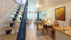 Un coin repas avec un escalier, des chaises en osier, une table avec deux verres à vin et un tableau accroché au mur.