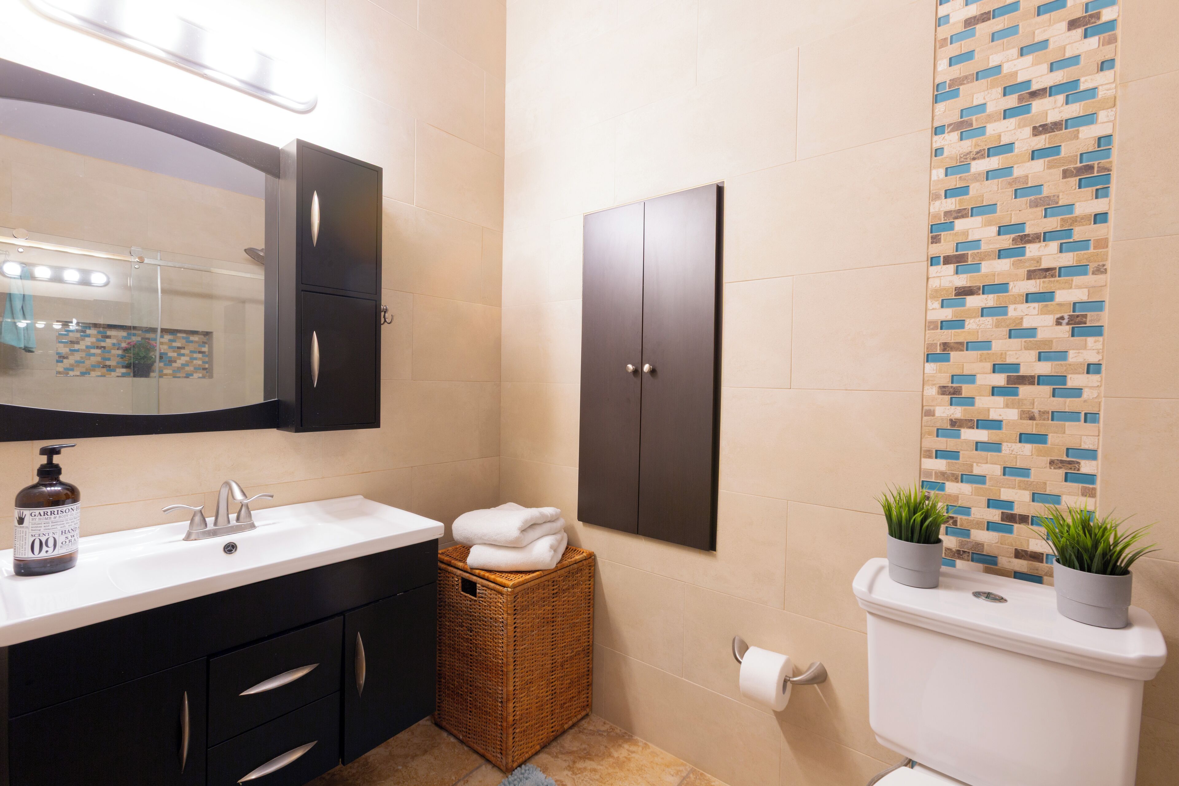 A bathroom with a white sink, dark cabinets, a wicker basket, and two potted plants on the toilet tank.