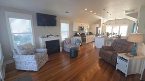 Uma sala de estar ampla com lareira, televisão e cozinha ao fundo.