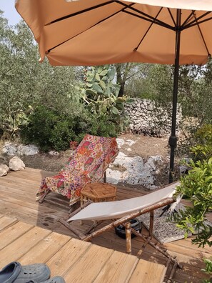 A wooden deck with two lounge chairs, one with a colorful patterned cover, and an umbrella providing shade.
