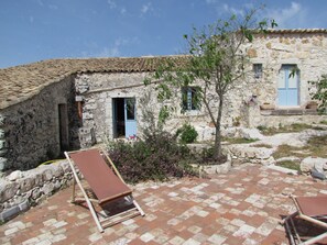 Un bâtiment en pierre avec un toit en tuiles, un petit jardin fleuri et deux chaises longues sur une terrasse dallée.