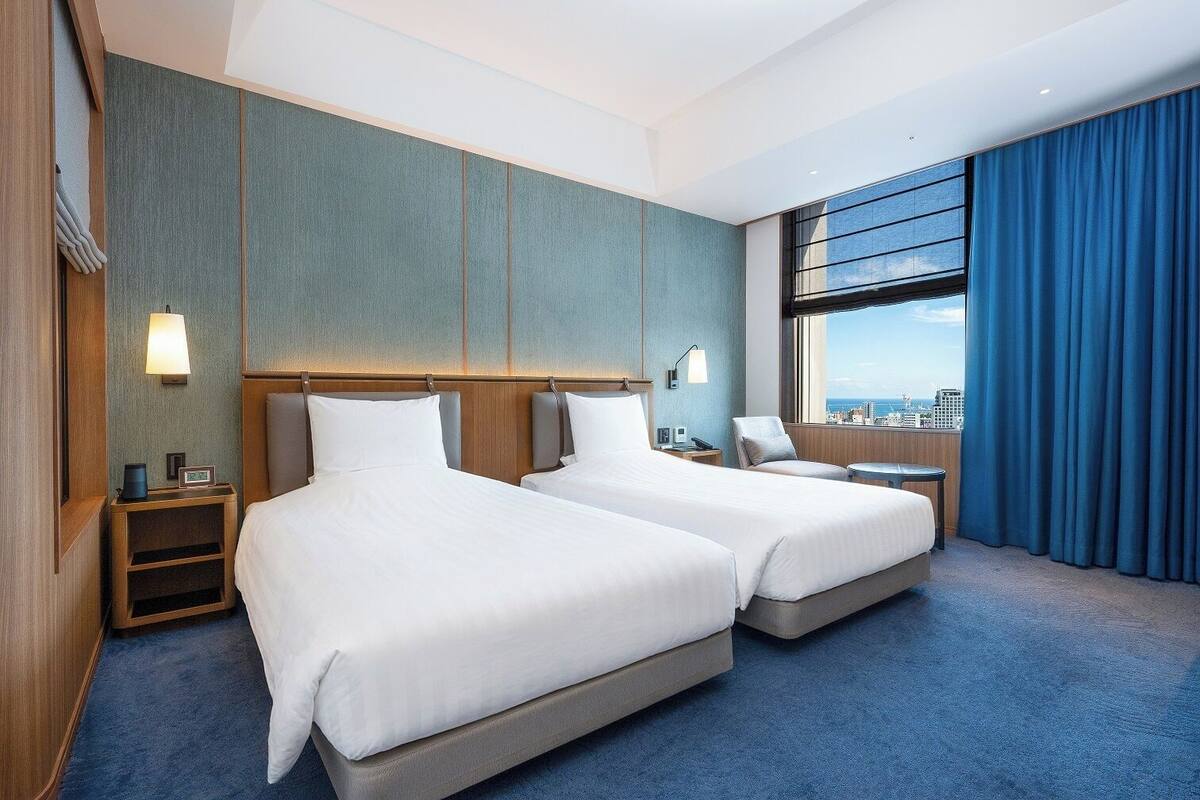 A hotel room with two beds, a view of the city, and a large window with curtains.