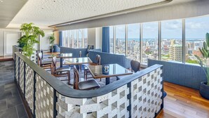 A modern bar area with a glass-top table, cushioned seating, and a city view through large windows. - Hotel Collective (Naha)